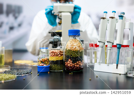 Scientist wearing blue gloves works with a microscope beside jars of beans, lentils and grains, with pipettes and test tubes in a food research laboratory. Scientist wearing blue gloves works with a microscope beside jars of beans, lentils and grains, with pipettes and test tubes in a food research laboratory. 142951059