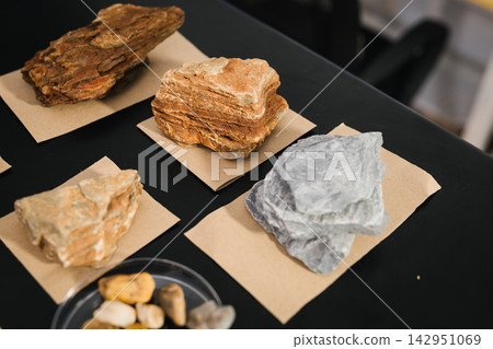 Close-up of different layered rock and mineral samples displayed on small paper cards on a black table during a geology exhibit or classroom lesson. Close-up of different layered rock and mineral samples displayed on small paper cards on a black table during a geology exhibit or classroom lesson. 142951069
