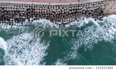 Aerial view of dramatic waves hitting concrete breakwater along a scenic coastal promenade, showcasing nature's power and human engineering. ocean, travel, and coastal infrastructure themes. Aerial view of dramatic waves hitting concrete breakwater along a scenic coastal promenade, showcasing nature's power and human engineering. ocean, travel, and coastal infrastructure themes. 142952900