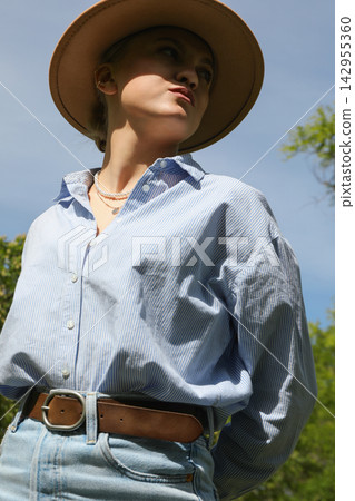 Woman poses outdoors wearing a blue shirt and hat under the sun Woman poses outdoors wearing a blue shirt and hat under the sun 142955360