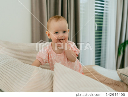 Adorable small child exploring hands while chewing fingers on couch in cozy living room. Concept of early childhood milestones, natural teething, and pediatric health Adorable small child exploring hands while chewing fingers on couch in cozy living room. Concept of early childhood milestones, natural teething, and pediatric health 142956400