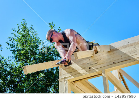 Roof truss installation by young worker using cordless drill to fasten rafter legs and timber trusses in wooden framework. Roof truss installation by young worker using cordless drill to fasten rafter legs and timber trusses in wooden framework. 142957690