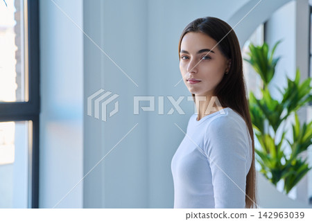 Portrait of serious young woman standing near window side view Portrait of serious young woman standing near window side view 142963039