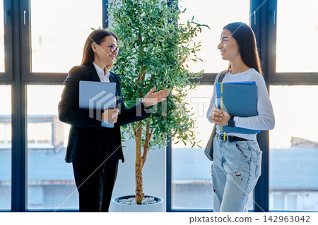 Teacher discussing lesson with student holding notebooks near window in modern office 142963042