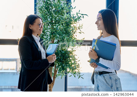 Teacher discussing lesson with student holding notebooks near window in modern office Teacher discussing lesson with student holding notebooks near window in modern office 142963044