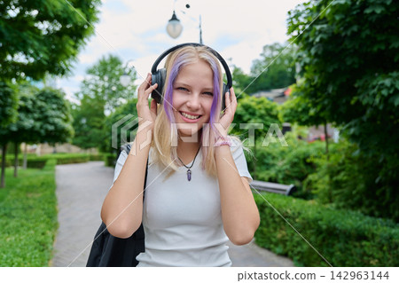 Smiling teenage girl listening to music in wireless headphones outdoors in park Smiling teenage girl listening to music in wireless headphones outdoors in park 142963144