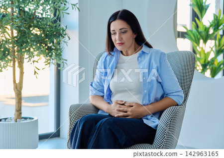 Middle aged woman holding stomach suffering from abdominal pain in armchair Middle aged woman holding stomach suffering from abdominal pain in armchair 142963165