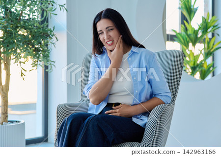 Middle aged woman touching her cheek suffering from severe toothache in armchair Middle aged woman touching her cheek suffering from severe toothache in armchair 142963166