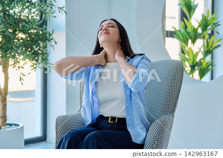 Middle aged woman touching her painful neck while sitting in armchair at home Middle aged woman touching her painful neck while sitting in armchair at home 142963167