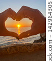 Hands making a heart shape around the setting sun over the ocean at golden hour. 142964305