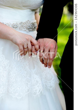 A close-up shot of a bride and groom holding hands, highlighting their matching gold wedding rings against the backdrop of a white lace bridal gown. A close-up shot of a bride and groom holding hands, highlighting their matching gold wedding rings against the backdrop of a white lace bridal gown. 142964306