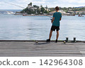 A man stands on a pier by the water, fishing. 142964308