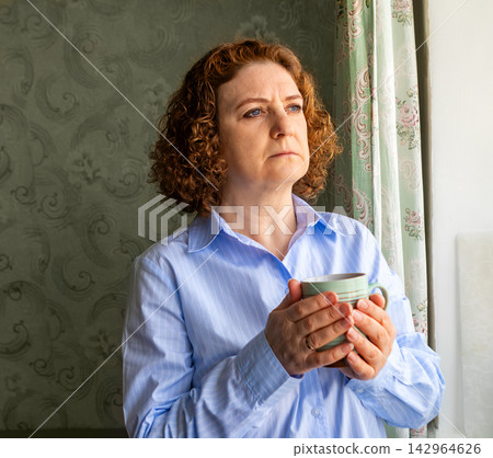Mature woman holding a mug and looking out the window in a thoughtful domestic portrait Mature woman holding a mug and looking out the window in a thoughtful domestic portrait 142964626