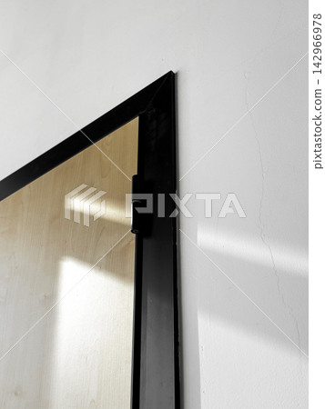 Closeup view of angle of wooden door on white wall. Door frame black, modern design. Sunlight casting shadows on wall. Intersection of light and shadow, room interior details Closeup view of angle of wooden door on white wall. Door frame black, modern design. Sunlight casting shadows on wall. Intersection of light and shadow, room interior details 142966978
