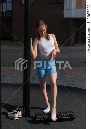 A Caucasian woman stretches her feet with a foam roller at a sports ground. 142967312