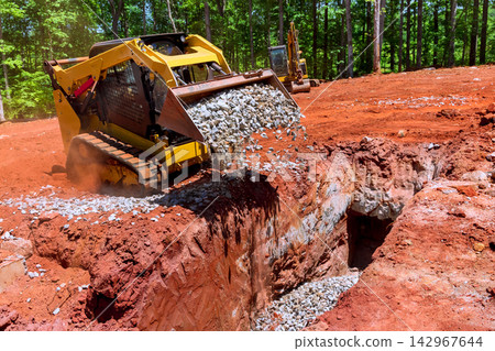 Heavy bulldozer machinery is busy filling trench with gravel at wooded construction site Heavy bulldozer machinery is busy filling trench with gravel at wooded construction site 142967644