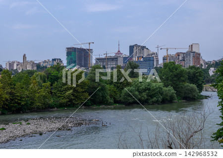 Scenic landscape photography of a wide river and a developing urban skyline with high-rises and construction cranes under an overcast sky 142968532