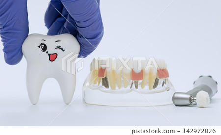 Overcoming dental fear concept with doctor hand holding a funny smiling tooth character near a clear acrylic prosthesis model and medical handpiece. Overcoming dental fear concept with doctor hand holding a funny smiling tooth character near a clear acrylic prosthesis model and medical handpiece. 142972026