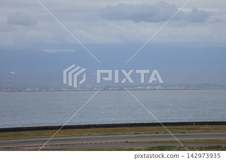 A landscape featuring an airport with a runway, the sea, an industrial complex with tanks and chimneys, and a cloudy sky. A landscape featuring an airport with a runway, the sea, an industrial complex with tanks and chimneys, and a cloudy sky. 142973935