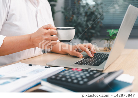 Business professional holding a coffee cup while using a laptop beside calculator and financial reports, representing office productivity, accounting, planning, finance, work break, Business professional holding a coffee cup while using a laptop beside calculator and financial reports, representing office productivity, accounting, planning, finance, work break, 142973947