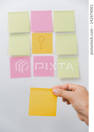 Hand holding a blank yellow sticky note in front of colorful notes with a question mark, representing decision making, uncertainty, problem solving, planning, priority, inquiry, Hand holding a blank yellow sticky note in front of colorful notes with a question mark, representing decision making, uncertainty, problem solving, planning, priority, inquiry, 142974001
