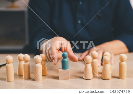 Hand highlights a teal wooden figure standing on a block among a team, representing talent recognition, leadership, promotion, candidate selection, career growth, hiring, and employee success. Hand highlights a teal wooden figure standing on a block among a team, representing talent recognition, leadership, promotion, candidate selection, career growth, hiring, and employee success. 142974759