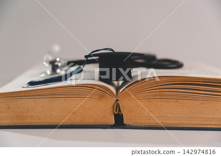 Graduation cap on an open book with stethoscope in the background, representing medical graduation, academic success, healthcare education, and the transition into a healthcare profession. Graduation cap on an open book with stethoscope in the background, representing medical graduation, academic success, healthcare education, and the transition into a healthcare profession. 142974816