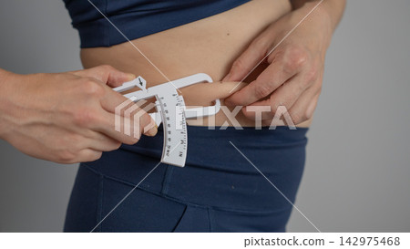 A woman measures the amount of subcutaneous fat on her abdomen using a caliper. A woman measures the amount of subcutaneous fat on her abdomen using a caliper. 142975468