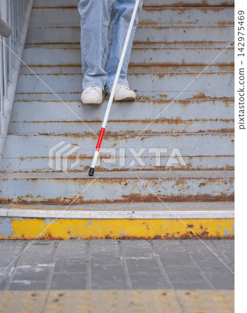 A blind woman descends a staircase. Close-up of her feet and a tactile stick. 142975469