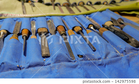 Set of woodcarving chisels and gouges arranged in blue fabric roll with wood shavings, worn wooden handles, metal blades, and shallow depth of field in workshop light Set of woodcarving chisels and gouges arranged in blue fabric roll with wood shavings, worn wooden handles, metal blades, and shallow depth of field in workshop light 142975598