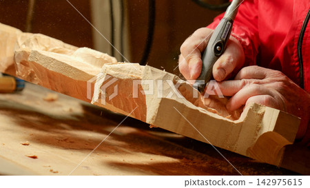 Hands carving softwood block with rotary tool, creating detailed relief texture and flying wood chips in warm workshop light, showing skilled artisan control and precise manual craft Hands carving softwood block with rotary tool, creating detailed relief texture and flying wood chips in warm workshop light, showing skilled artisan control and precise manual craft 142975615