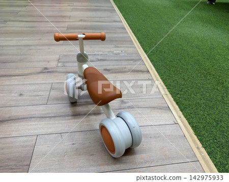 Three wheel toddler balance bike with orange handlebars on wooden floor in playroom Three wheel toddler balance bike with orange handlebars on wooden floor in playroom 142975933