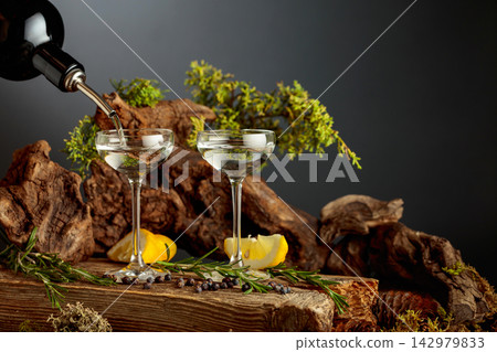 Gin poured from a bottle into a glass, set among juniper branches, weathered driftwood and forest moss. Gin poured from a bottle into a glass, set among juniper branches, weathered driftwood and forest moss. 142979833