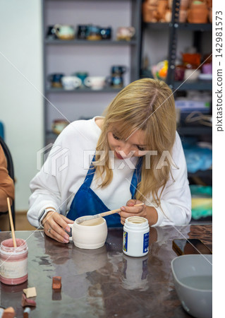 Young woman applying glaze to handmade ceramic mug with brush in pottery workshop. Pottery glazing process represents creativity and artisan craftsmanship Young woman applying glaze to handmade ceramic mug with brush in pottery workshop. Pottery glazing process represents creativity and artisan craftsmanship 142981573