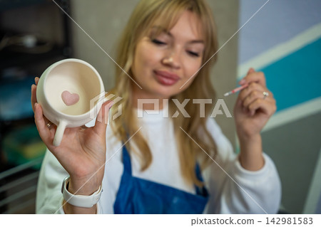 Young woman showing handmade ceramic mug with painted heart after pottery class. Finished craft represents creativity, self expression and satisfaction with handmade work Young woman showing handmade ceramic mug with painted heart after pottery class. Finished craft represents creativity, self expression and satisfaction with handmade work 142981583