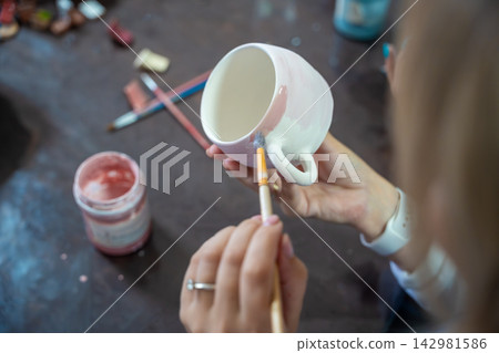 Woman painting pink glaze on handmade ceramic mug with brush in pottery workshop. Pottery decoration represents creativity and learning a craft as a hobby Woman painting pink glaze on handmade ceramic mug with brush in pottery workshop. Pottery decoration represents creativity and learning a craft as a hobby 142981586
