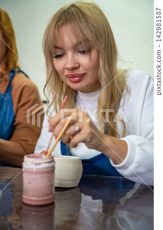 Woman dipping paintbrush into ceramic glaze while decorating handmade mug. Preparing brush with glaze represents detailed pottery painting process and artisan craft Woman dipping paintbrush into ceramic glaze while decorating handmade mug. Preparing brush with glaze represents detailed pottery painting process and artisan craft 142981587