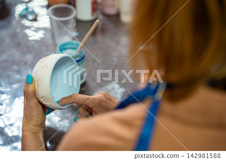 View of woman hands smoothing glaze on handmade ceramic mug with finger after painting. Removing air bubbles and imperfections represents finishing process in pottery craft 142981588