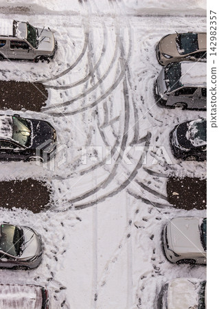 An overhead view of a snow-covered parking lot on a day of severe cold snap, showing the patterns of tire tracks and footprints left on the ground alongside the cars. An overhead view of a snow-covered parking lot on a day of severe cold snap, showing the patterns of tire tracks and footprints left on the ground alongside the cars. 142982357