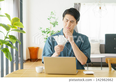 一位年輕的日本男子在家裡的桌子上,一邊查看收據,一邊用電腦記錄家庭預算。 一位年輕的日本男子在家裡的桌子上,一邊查看收據,一邊用電腦記錄家庭預算。 142982662