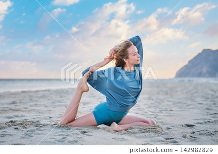 Woman performs Eka Pada Rajakapotasana on sandy beach at sunset Woman performs Eka Pada Rajakapotasana on sandy beach at sunset 142982829
