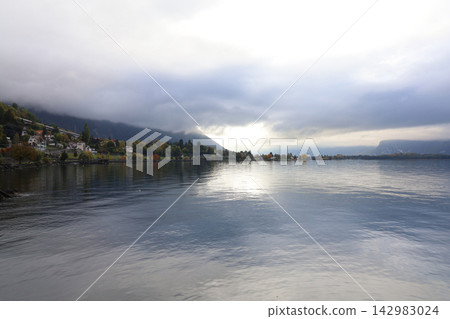 Moody autumn lake landscape with dramatic storm clouds, sunlight breaking through, misty mountain and lakeside village with colorful fall trees Moody autumn lake landscape with dramatic storm clouds, sunlight breaking through, misty mountain and lakeside village with colorful fall trees 142983024