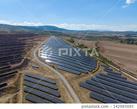 Solar panels line rural hillside rows, forming large power array beside dirt roads. Bright sunlight reflects from modules, and renewable energy site spans open fields under clear sky. Solar panels line rural hillside rows, forming large power array beside dirt roads. Bright sunlight reflects from modules, and renewable energy site spans open fields under clear sky. 142983085