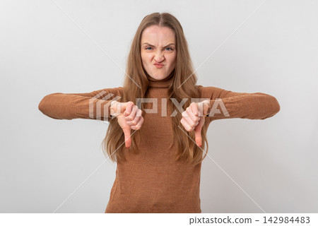 Young woman showing both hands pointing thumbs down while grimacing, expressing strong disapproval, dislike, and negative feedback on a plain light background Young woman showing both hands pointing thumbs down while grimacing, expressing strong disapproval, dislike, and negative feedback on a plain light background 142984483