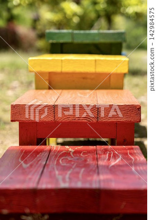 A charming scene unfolds outdoors amidst lush greenery, with small wooden chairs in a variety of colors—red, yellow, brown, and more—lined up in a single row. A charming scene unfolds outdoors amidst lush greenery, with small wooden chairs in a variety of colors—red, yellow, brown, and more—lined up in a single row. 142984575