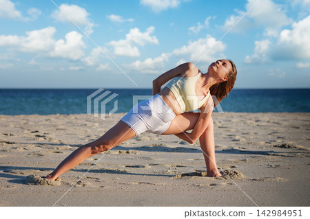 Woman performs Baddha Trikonasana on beach at sunset in wide stance Woman performs Baddha Trikonasana on beach at sunset in wide stance 142984951
