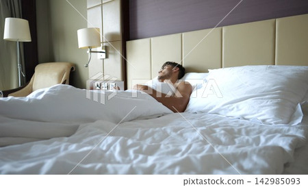 Young man sleeping peacefully in large bed in modern hotel room or home interior. Handsome guy resting under soft linen in cozy indoor atmosphere with natural comfort and relaxation Young man sleeping peacefully in large bed in modern hotel room or home interior. Handsome guy resting under soft linen in cozy indoor atmosphere with natural comfort and relaxation 142985093