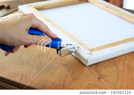 Person Removing Staples from Canvas Stretcher Frame with Blue Tool. Art Framing Refurbishment Concept Person Removing Staples from Canvas Stretcher Frame with Blue Tool. Art Framing Refurbishment Concept 142985114