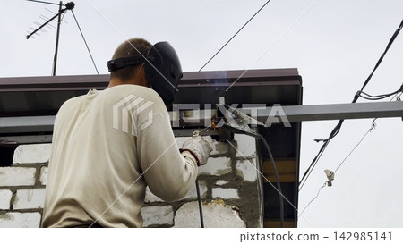 Manual laborer in protective mask welds iron detail at construction. Professional young repairman with welding machine working at the site. Male welder operating with metal on house. Close up Manual laborer in protective mask welds iron detail at construction. Professional young repairman with welding machine working at the site. Male welder operating with metal on house. Close up 142985141