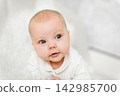 Baby 5-6 months old in white clothes lies on tummy at home and looks at camera, bright portrait with blue eyes 142985700